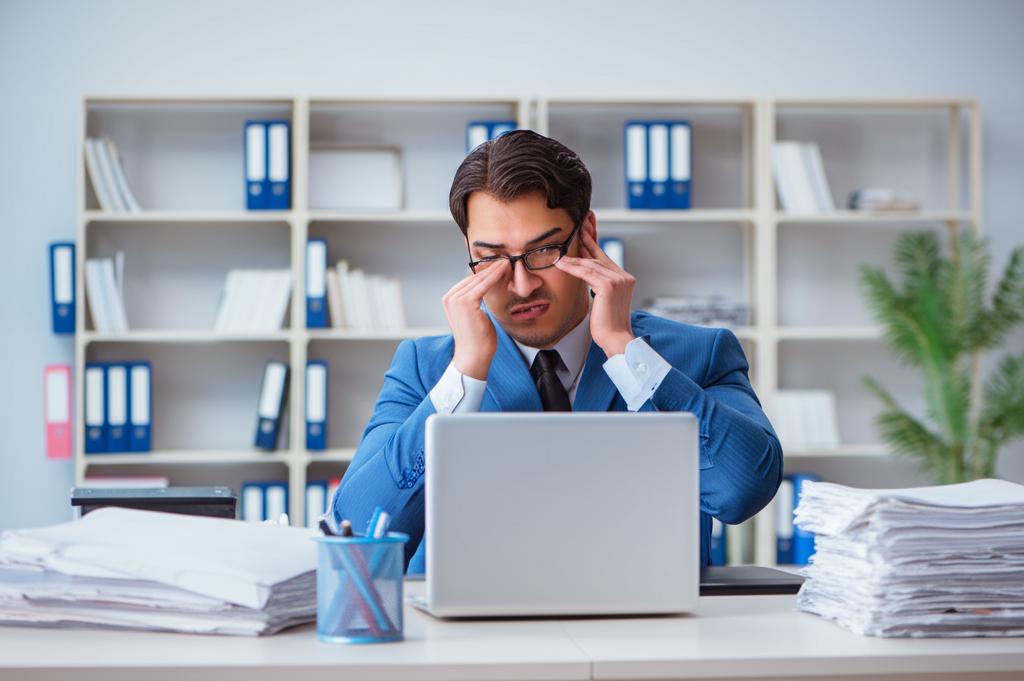 A stressed businessman sitting at his desk, overwhelmed with paperwork, looking exhausted.