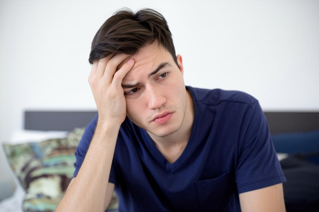 A young man looking stressed and disappointed, sitting alone, with a blurred background suggesting a bedroom.