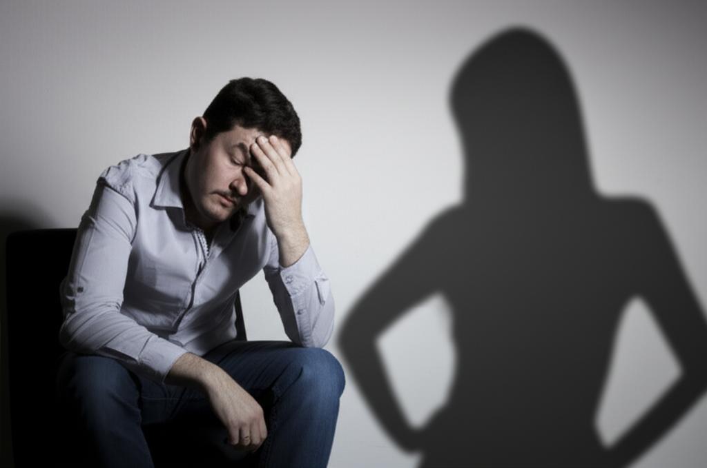 A man looking tired and stressed, sitting alone, with a shadow of a disappointed woman in the background, conveying psychological burden and relationship strain.
