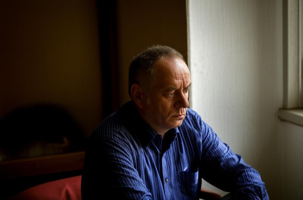 AI prompt: A man sitting alone, looking worried and deep in thought, perhaps reflecting on past experiences, in a slightly dim room.